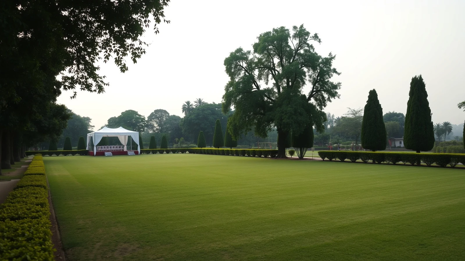Vast empty manicured lawn wedding venue near Delhi in soft evening light, with a white marquee set back among mature trees